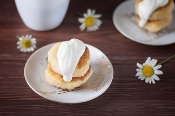 Gourmet breakfast - homemade pastries from cottage cheese, cheesecakes, cottage cheese pancakes with sour cream in a white plate on a dark wooden background.
