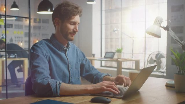 Handsome Creative Male in Jeans Shirt Works on a Laptop in His Sunny Loft Office. He Looks Cool and Smart, He Has a Beard. Colleague Works with Storyboard on the Background. Office Has a Big Window.