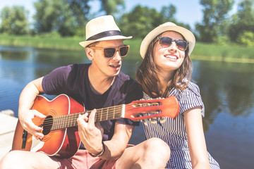 Beautiful fashionable couple with acoustic guitar against lake or river in the background. Summer and relationship concept.