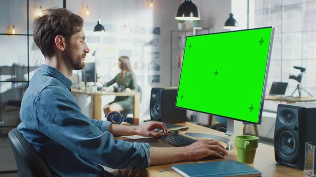 Male Video Editor with Beard and Jeans Shirt Works on His Personal Computer with Big Green Screen Mock Up Display. He Works in a Cool Office Loft. Other Female Creative Colleague Walks in Background.