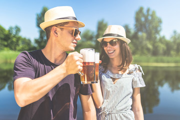 Young fashionable couple holding beer glasses against beautiful nature. Summer party concept.