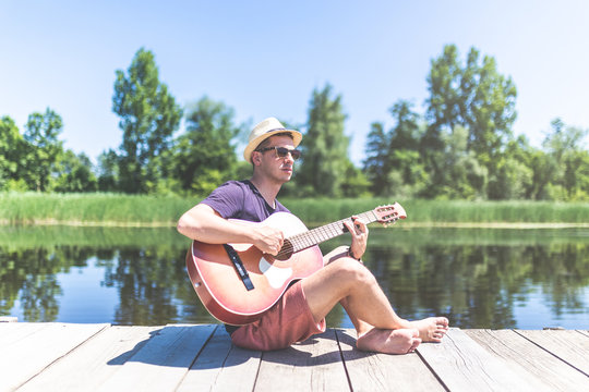 Young hipster guy holding acoustic guitar while sitting on dock in nature. Summer and travel concept.