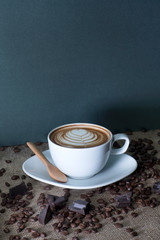 A cup of fresh coffee with a flower formed cream and a wooden spoon on the gunny sack table with coffee beans and chocolate