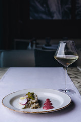 Restaurant delicacy dish on a purple tablecloth with glass of white wine. White plate with herring, gherkins, red onions and quail eggs. Farshmak with beetroot sauce.