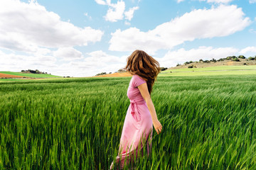 A long-haired woman in a pink dress in a green field of wheat on the background of the mountains. Spanish landscape. Beautiful agriculture sunset landscape. Sunrise sky. Green farm.