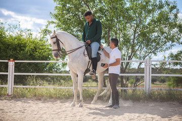 Young man teaching black guy to ride horse in paddock on cloudy day on ranch