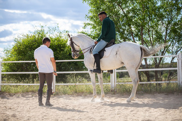Young man teaching black guy to ride horse in paddock on cloudy day on ranch