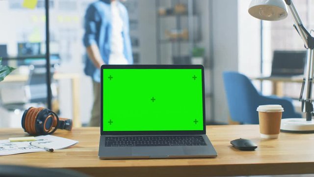 Laptop on the Desk in the Office Shows Green Mock-up Screen. In the Background Bright Modern Open Space Creative Office HUB with Professional Working