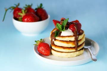 Syrniki served with sour cream, strawberry and melted chocolate. White plate, blue table, high resolution