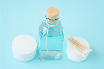 Face cream in white jar, face tonic or makeup remover in a glass bottle, cotton pads and wooden cotton buds on pastel blue background. Hygiene supplies, beauty tools and skin care. Top view.
