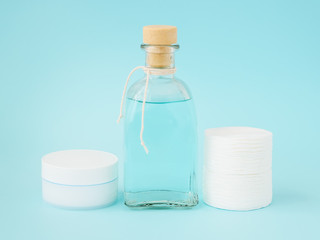 Fototapeta na wymiar Face cream in white jar, tonic for face skin or makeup remover in a glass bottle and cotton pads on pastel blue background. Hygiene supplies, beauty tools and skin care.
