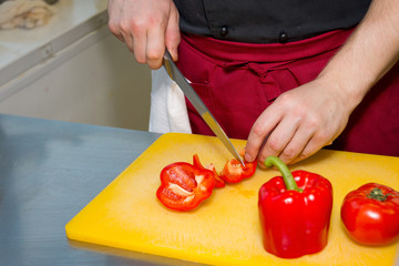 Tough guy cutting paprika at kitchen. Chef's hands with knife cutting red paprika on cutting board for salad. red paprika on yellow apron. How to cut paprika. bell pepper, bulgarian pepper