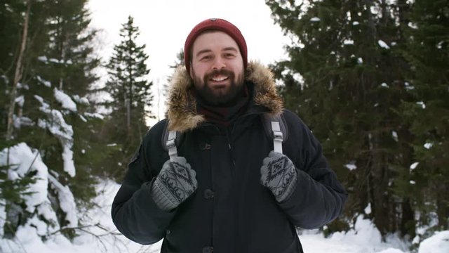 Portrait shot with handheld tracking of happy bearded man with backpack smiling and posing for camera in forest on winter day