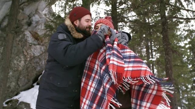 Handheld tracking shot of affectionate young man with beard wrapping blanket around his shivering girlfriend while hiking in mountains on cold winter day. He is smiling and giving her kiss on lips