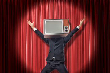 Happy businessman with vintage tv set instead of head raising arms on red stage curtains background