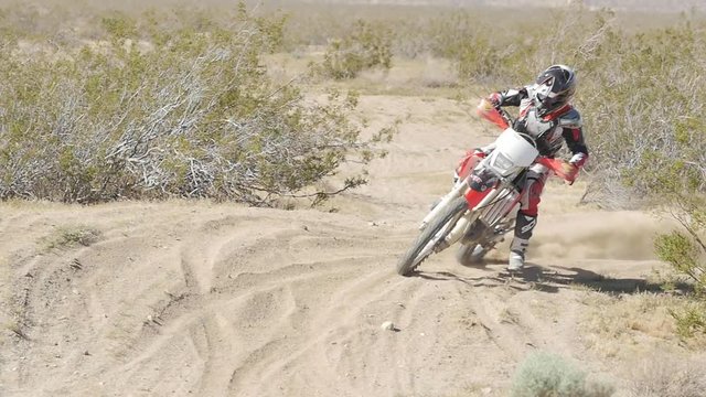 SLOW MOTION: A dirt biker rides his CRF Honda through the desert and whips around a berm. Comes around the corner and takes off.