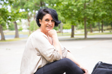 Happy confident woman relaxing in park. Middle aged black haired lady sitting against green trees and smiling at camera. Woman outdoors concept