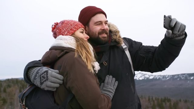 Handheld shot of happy young couple with backpacks standing on top of mountain on cold winter day and taking selfie on mobile phone