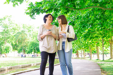 Naklejka premium Positive young woman and her mother chatting and walking in park. Middle-aged lady and her daughter walking on boardwalk and relaxing with trees in background. Family and nature concept. Front view.