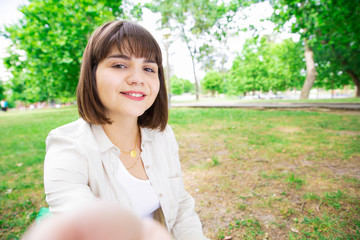Smiling pretty woman taking selfie photo and sitting on lawn. Young lady holding gadget which is out of view and sitting on ground with trees in background. Selfie and nature concept. Front view.