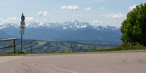 Fototapeta na wymiar PEISSENBERG, BAVARIA / GERMANY - June 2, 2019: Panorama view of Wetterstein mountain range. In the middle Zugspitze, Germanys highest mountain, The mountains are framed by a sign post and bushes.
