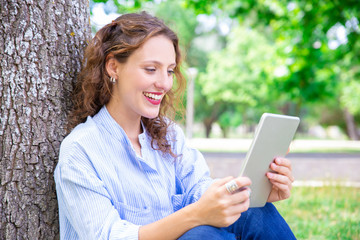 Fototapeta premium Happy young woman talking via telecommunication app on tablet. Jolly curly-haired girl chatting online in park. Video chat concept