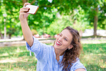 Jolly charming lady winking while taking selfie in park. Happy young woman with wavy hair resting...