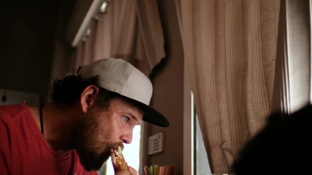 blue-eyed hipster guy with a beard and a cap snapback eating a sandwich (burger)  in a street cafe eating a sandwich (burger) in a street cafe. 4k. 4k video