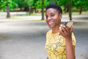 Smiling Black woman listening audio message on smartphone. Beautiful positive African-American young woman recording message for friend when walking in park. Communication concept