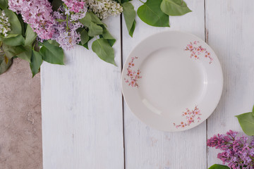 A plate and a decor of flowers on a background of white-painted wooden boards. Vintage background with lilac flowers and a place under the text. View from above. Cutlery.