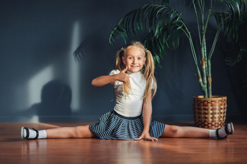 Beautiful little blonde girl with hair gathered in tails, white t-shirt, white socks and gray skirt sitting on twine at home, shows the thumb, looking at camera and smiles.