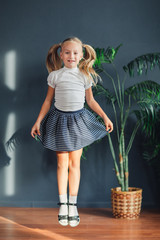 8 years old Beautiful little blonde girl with hair gathered in tails, white t-shirt, white socks and gray skirt jumping in a child room at home, still life photo.