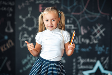 Cute girl in school uniform with colored pencils. Go to school for the first time. Girl indoors of the class room with blackboard on a background shows the thumb. Back to school.