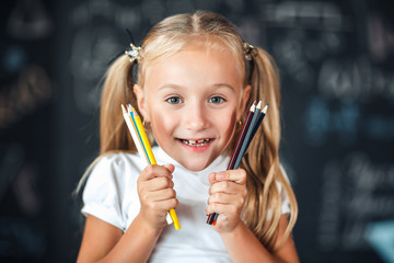 Back to school! Portrait a little smiles girl stands with girl holding colored pencils near the face against chalkboard with school formulas at school.