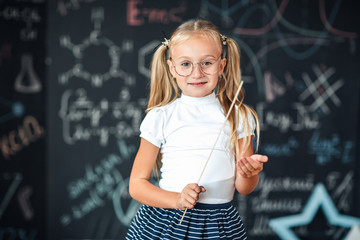 Cute schoolgirl with glasses and ruler looking like a strict teacher raised her pointer to draw attention against chalkboard with school formulas. Educational concept. School concept. Back to School