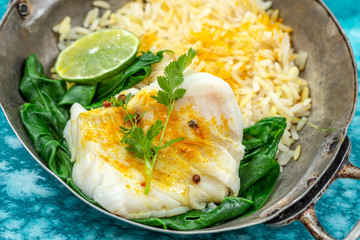 Fototapeta na wymiar Cod fillet with rice and spinach garnish with curry, in vintage pan on blue marbled wooden background