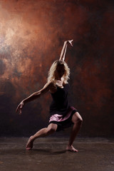 Fototapeta na wymiar Photo of young dancing blonde with raised arms in black dress on brown background