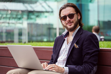 Photo of young brunet businessman with laptop sitting on wooden bench in city center on summer day