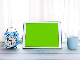 Green screen tablet computer, blue alarm clock and coffee mug on blue wooden table.