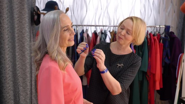 Portrait of two women in shop. Seller consultant offers an elderly elegant grey-haired woman to try on earrings in store.
