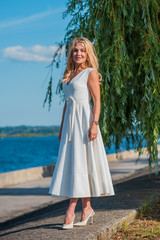 Fototapeta na wymiar Elegant blonde hair woman in white midi dress at shore. Concept oа white code, fashionable lady, pretty dress and clothes 