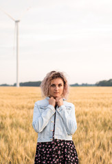 young tanned curly haired woman in denim jacket and dress on the field of ripened wheat, sunset time