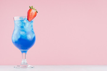 Tropical fresh blue drink with curacao, ice cubes, strawberry in exquisite misted wineglass on pastel pink wall, white table.
