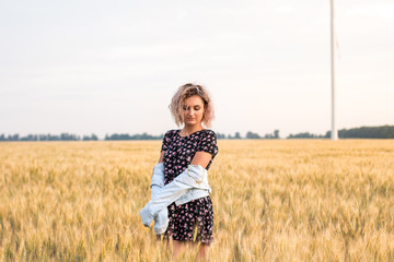young tanned curly haired woman in denim jacket and dress on the field of ripened wheat, sunset time