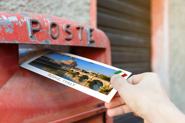 Male hand is putting a postcard in a typical red postbox in Rome, Italy. The postcard shows a famous sights of Rome.