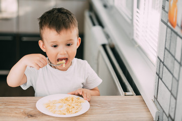 a child in the kitchen during the day eating pasta in a spiral in a white t-shirt