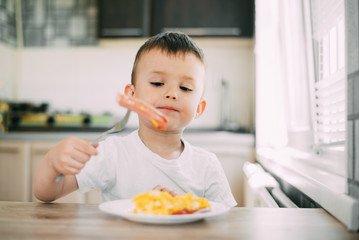 a child in a t-shirt in the kitchen eating a sausage and an omelet with a fork is very appetizing