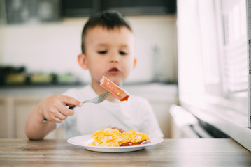 a child in a t-shirt in the kitchen eating a sausage and an omelet with a fork is very appetizing