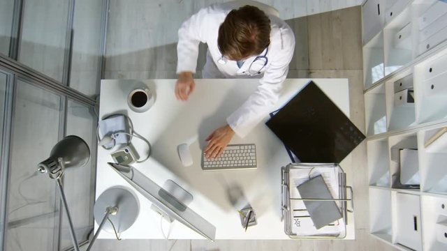 Top view shot of male doctor in white coat sitting at his desk in office and drinking coffee while working on computer