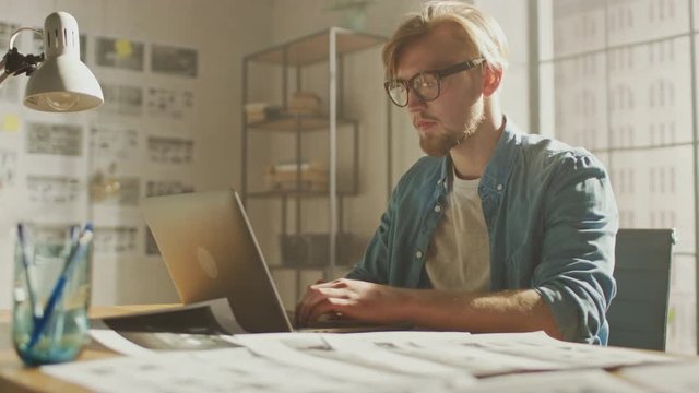 Creative Designer Uses on a Laptop Sitting at His Desk in the Modern Studio, Working on a Storyboard, Project Sketches and Concepts Hanging on the Walls. Video Editing, Application UI or Game Plot